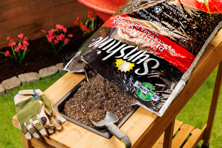 Bag of SunGro potting mix spilled into a wooden planter with gardening gloves, trowel and pink flowers in background