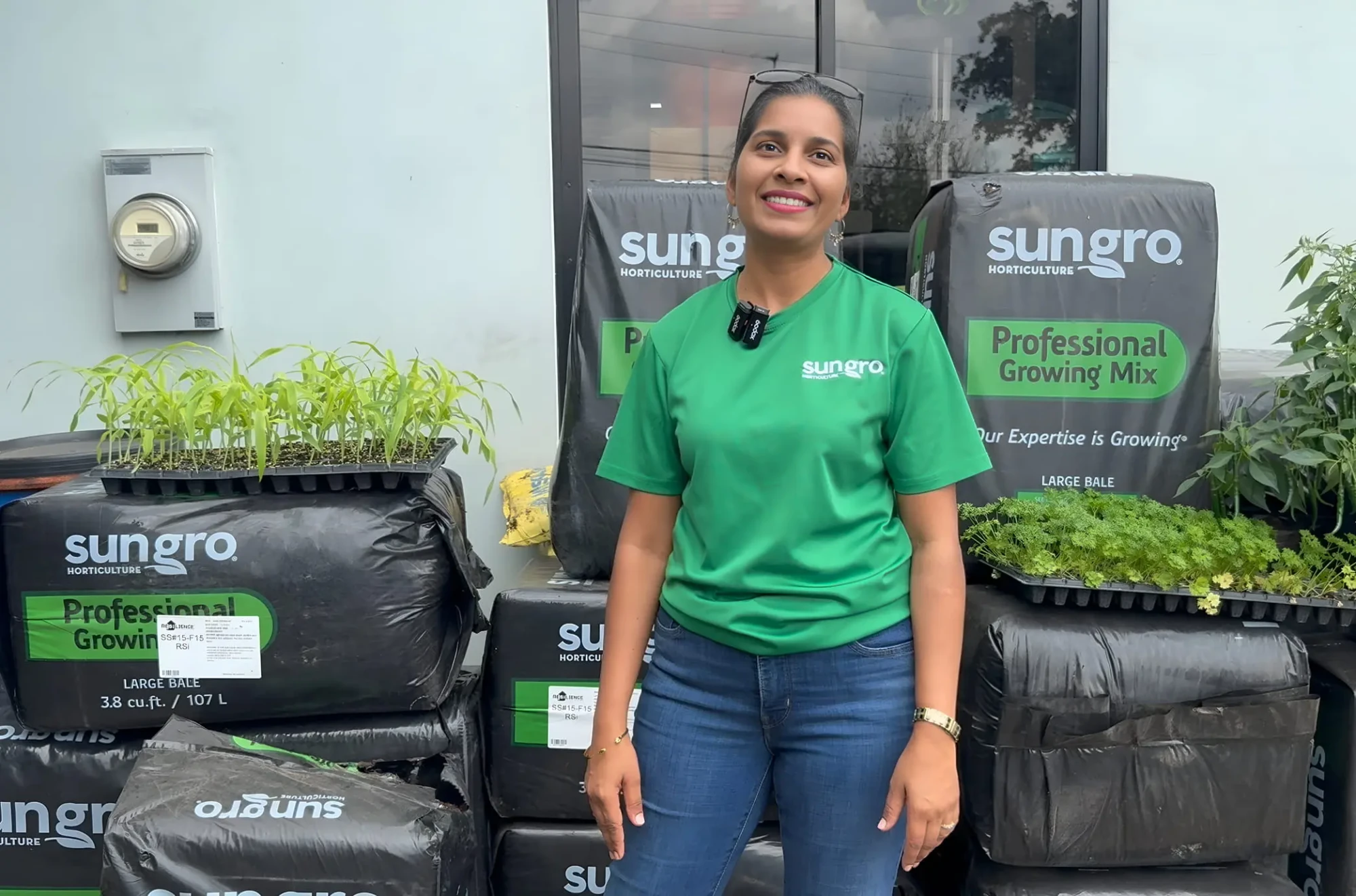 Smiling Sungro employee in green shirt stands beside stacked Sungro growing mix bales and trays of seedlings at nursery